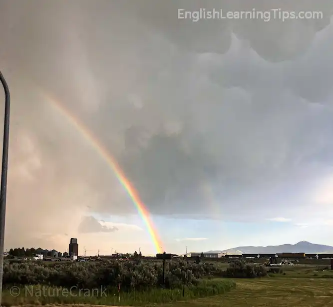 A vibrant rainbow arching across a dark, stormy sky over a rural landscape