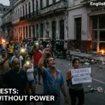 A crowd of people protesting on a dark Cuban street at night with flashlights and a sign that reads "¡Queremos luz, no más apagones!" near a damaged building.